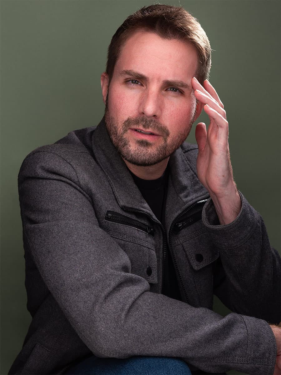 Scott Squire in a grey jacket, hand at his temple, against a green backdrop