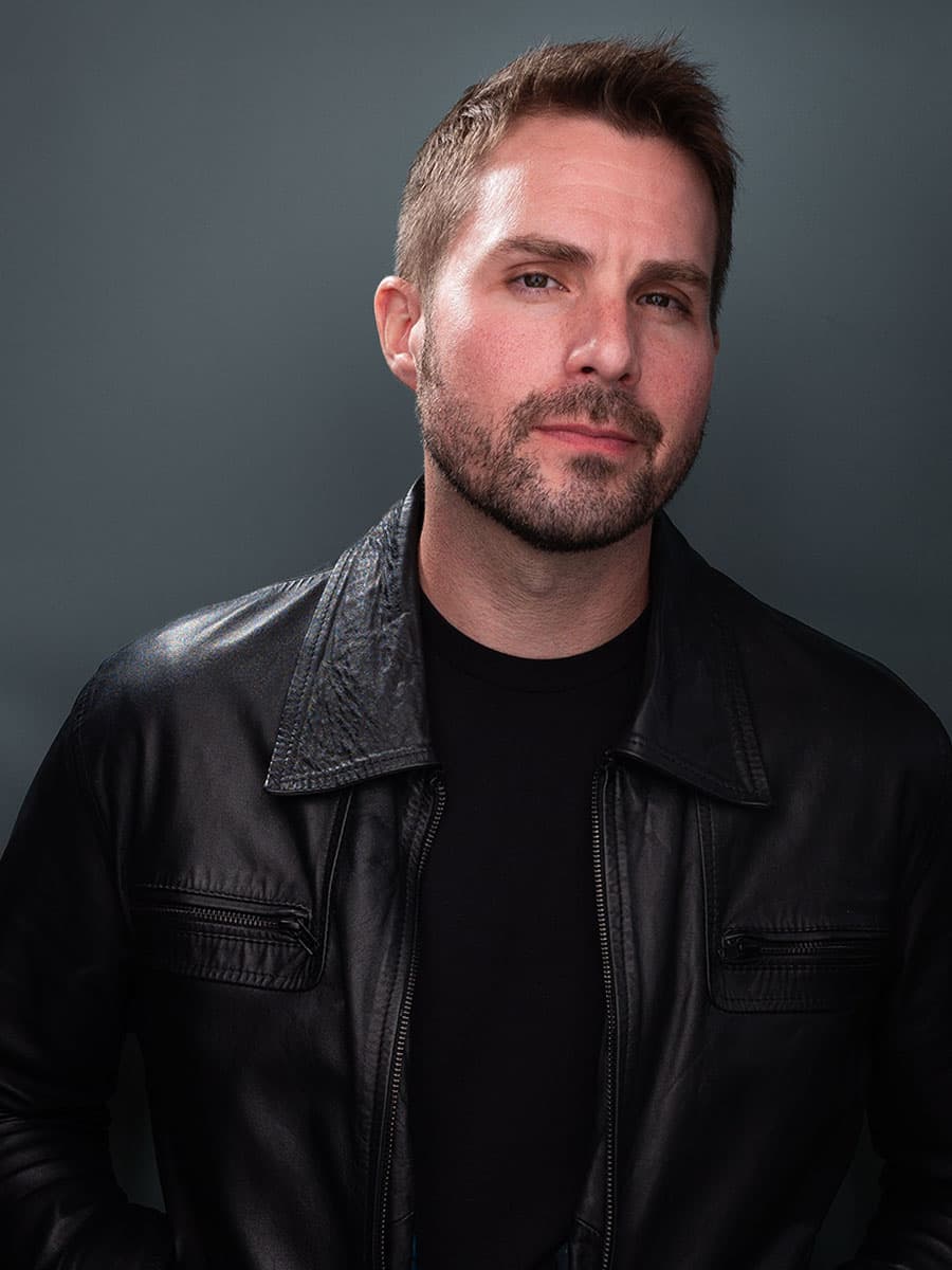 Scott Squire in a black leather jacket, head tilted, against a grey backdrop
